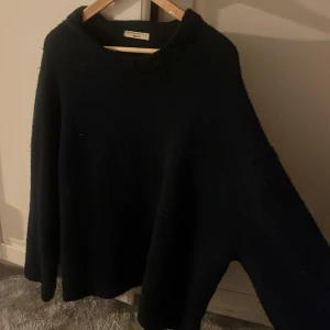 Svart stickad tröja från Gina Tricot - En svart, oversized stickad tröja från Gina Tricot i storlek L. Tröjan har rund hals och långa ärmar, perfekt för att hålla sig varm under kyliga dagar. Mjuk och skön kvalitet som känns lyxig mot huden. Enkel och stilren design som funkar till många olika looks.