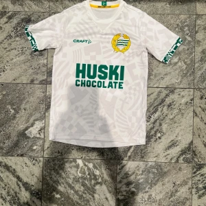 Vit Hammarby fotbollströja Craft junior - Snygg vit Hammarby fotbollströja från Craft i juniorstorlek. Tröjan har grönt tryck med HUSKI CHOCOLATE på bröstet, klubbmärke i gult och grönt, samt diskret geometriskt mönster. Kortärmad med gröna detaljer på ärmslut och texten 'UNDER GRÖNVIT FLAGG' i nacken. Perfekt för match eller träning.