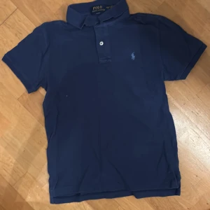 Marinblå Polo Ralph Lauren t-shirt - Snygg marinblå t-shirt från Polo Ralph Lauren med klassisk pikékrage och knappslå. Slim fit-modell med korta ärmar och den ikoniska broderade loggan på bröstet. Tillverkad i mjuk bomull som är skön mot huden. Perfekt för en clean och stilren look.