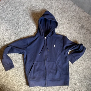 Marinblå hoodie från Polo Ralph Lauren - Snygg marinblå hoodie från Polo Ralph Lauren med dragkedja och huva. Tröjan har långa ärmar, två fickor fram och den klassiska vita Polo-loggan på bröstet. Tillverkad i mjuk bomull och polyester, perfekt för chill dagar eller när du vill ha en clean look.