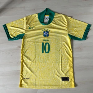 Brasilien Neymar Jr. 10 Nike fotbollströja - Gul Brasilien fotbollströja från Nike med gröna detaljer och nummer 10 på både fram- och baksidan. Neymar Jr. tryck på ryggen, officiellt CBF-emblem och Nike-logga. Tillverkad i lätt, ventilerande Dri-Fit material med subtilt mönster över hela tröjan. Perfekt för dig som älskar fotboll och Brasilien. Som ny