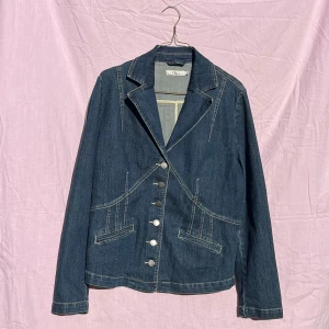 Mörkblå jeanskavaj från Olé Jeans - Snygg mörkblå jeanskavaj från Olé Jeans med klassisk krage och silverfärgade knappar framtill. Jackan har markerade sömmar, två fickor och långa ärmar med knappdetaljer. Materialet är en mix av bomull, polyester och elastan för skön passform.