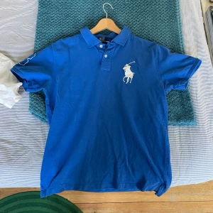 Blå Polo Ralph Lauren t-shirt - Snygg blå t-shirt från Polo Ralph Lauren med klassisk polokrage och två knappar framtill. Stor vit broderad logga på bröstet och cool detalj på ärmen. Tillverkad i mjuk bomull, perfekt för dig som gillar stilren och sportig look.