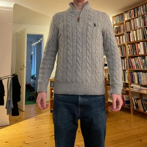 Grå stickad half zip-tröja Polo Ralph Lauren - Säljer en grå stickad tröja från Polo Ralph Lauren med half zip och klassisk kabelstickad design. Tröjan har lång ärm, ribbade muddar och krage, samt den ikoniska logon broderad på bröstet. Perfekt för dig som gillar stilren och tidlös look.