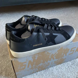Golden Goose Super-Star svarta sneakers - Svarta Golden Goose Super-Star sneakers med ikonisk stjärndetalj i mocka på sidan, svarta snören och vit platt sula. Skorna har detaljer i både skinn och mocka, samt guldfärgade tryck på sidan och plös. Snygg och stilren design som sticker ut.
