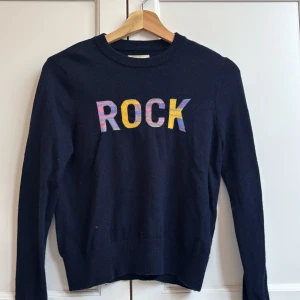 Mörkblå kashmirtröja Zadig & Voltaire - Snygg mörkblå kashmirtröja från Zadig & Voltaire med färgglad 'ROCK'-text framtill. Tröjan har rund hals, långa ärmar och ribbade muddar. Perfekt för dig som gillar coola statement-plagg och mjuka material.