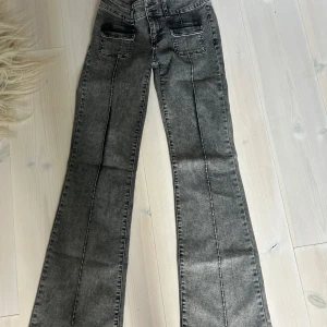 Bootcut jeans i grå från Nelly, EU 36 - Snygga grå bootcut jeans från Nelly med låg midja och coola fickdetaljer framtill. De har aldrig kommit till användning och är i ett nytt skick. Jeansen har markerade sömmar längs benen och är tillverkade i stretchigt material med bomull, polyester och elastan. Perfekt för dig som gillar en trendig och bekväm look.