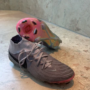 Nike Phantom grå fotbollsskor - Nike Phantom fotbollsskor i grått med stickad ovandel och svart Swoosh på sidan. Skorna har snörning, rund tå och en hög stickad krage för extra stöd. Sulan är skimrande med rosa och guldtoner, och insidan har en röd detalj med Phantom-logga. Perfekt för dig som vill sticka ut på planen.