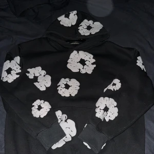 Denim tears  - Svart hoodie från Denim Tears med unika vita grafiska tryck över hela tröjan, inklusive huvan och ärmarna. Tröjan har huva och ribbade muddar. Perfekt för dig som gillar streetwear med en twist och vill sticka ut med cool design.