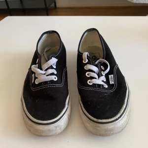 Svarta Vans sneakers med vit sula - Klassiska svarta Vans sneakers med vit platt sula och vita snören. Skorna har rund tå och tydliga vita sömmar som kontrast mot det svarta bomullstyget. Vans-logga på sidan och röd logga bak på hälen. Perfekt för dig som gillar enkel och tidlös streetstil.
