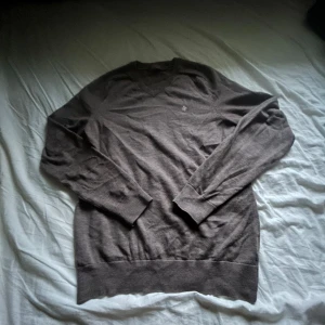 Brun v-ringad stickad tröja - Snygg brun stickad tröja från morris med v-ringad hals och diskret logga på bröstet. Tröjan har långa ärmar och ribbade muddar vid ärmslut och nederkant. Perfekt för dig som gillar klassisk stil med en modern touch.