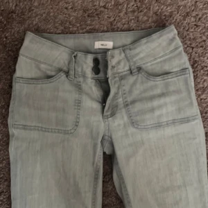 Ljusgrå bootcut jeans från Nelly - Snygga ljusgrå jeans från Nelly med bootcut passform och hög midja. Framtill finns två stora fickor och dubbla knappar i midjan, baktill har byxorna fickor med lock och knapp. 