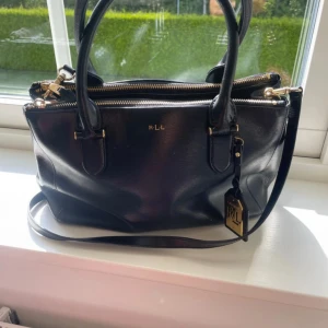 Svart handväska från Ralph Lauren - Snygg svart handväska från Ralph Lauren med guldfärgade detaljer och logotypen RLL på framsidan. Väskan har både handtag och axelrem, flera fack med dragkedja och en exklusiv tag i guld. Tillverkad i skinn med stilren och klassisk form. Ny pris ca 3000kr