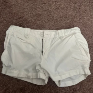 Vita midwaist shorts med knappar - Snygga vita shorts med midwaist passform och klassiska detaljer som knappar och dragkedja framtill. De har fickor både fram och bak samt breda hällor för bälte. Perfekta för varma dagar och har en stilren look som funkar till många outfits.