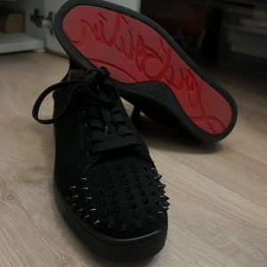 Christian Louboutin svarta sneakers med nitar - Svarta sneakers från Christian Louboutin med ikoniska röda sulor och svarta nitar på tån. Skorna har snörning och en rund tå, tillverkade i mocka och skinn. Perfekt för dig som vill sticka ut med edgy detaljer och lyxig vibe. Inget org medföljer så endast skorna! Buda gärna