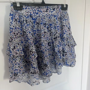 Blommig blå volangkjol från Zadig & Voltaire - Supersöt kort kjol från Zadig & Voltaire med blå botten och blommigt mönster i vitt, rosa och lite svart. Kjolen har flera volanger och ett elastiskt midjeband som gör den bekväm och lätt att bära. Perfekt för dig som gillar färg och detaljer!