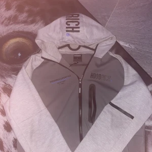 Grå hoodie från Hoodrich med tryck - Säljer en äkta grå hoodie från Hoodrich i storlek L. Tröjan har svart tryck på både fram- och baksidan samt på huvan. Den har dragkedja framtill och praktiska fickor med svarta detaljer. Perfekt för dig som gillar streetwear och snygga loggor.