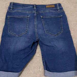 Blå jeansshorts med uppvik - Säljer ett par klassiska blå jeansshorts från Coul med snygga uppvikta benslut. Shortsen har fem fickor, bälteshällor och knappstängning framtill. Perfekt för dig som gillar en enkel och tidlös stil. Passar dig som vill ha bekväma shorts med normal passform.