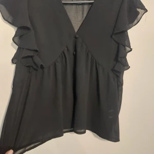 Svart blus med volangärmar - Säljer en svart blus med v-ringad hals och snygga volangdetaljer på ärmarna. Blusen är gjord i ett lätt och transparent material som ger en luftig känsla. Perfekt för dig som gillar en stilren look med lite extra edge. Passar till både jeans och kjol.