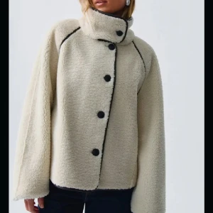 Beige teddyjacka med svarta detaljer - Säljer en beige teddyjacka med svarta knappar och svarta sömmar. Jackan har hög krage med knappstängning och är långärmad med en loose passform. Perfekt för kalla dagar och riktigt mysig att bära.