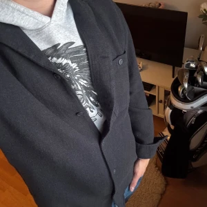 Svart overshirt från Dobber, M - Snygg svart overshirt från Dobber i storlek M. Jackan har klassisk krage, knäppning med svarta knappar och en bröstficka. Materialet känns mjukt och har en stilren look som funkar till jeans och hoodie. Perfekt för dig som gillar enkel och clean stil.
