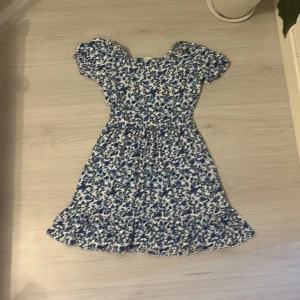 Blommig kortklänning från Lindex - Söt kortklänning från Lindex med blått och vitt blommigt mönster. Klänningen har puffiga korta ärmar, markerad midja och volangkant nedtill. Perfekt för dig som gillar en feminin och fräsch stil. Materialet är mjukt och lätt, troligen bomull eller polyester.