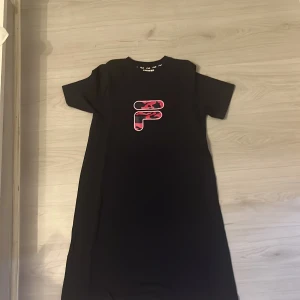 Svart kortklänning från FILA - Svart kortklänning från FILA med rosa och röd logga framtill i ett coolt camouflage-mönster. Klänningen har rund hals, korta ärmar och en rak, loose passform. Perfekt för dig som gillar sportiga och stilrena plagg med en twist.