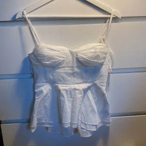 Vit korsett-topp från Zara - Supersnygg vit korsett-topp från Zara med figurnära passform och tunna axelband. Toppen har markerade kupor och en lätt utsvängd nederdel som ger en feminin siluett. Perfekt att styla med jeans eller kjol för en trendig look.