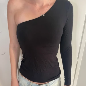 Svart oneshoulder topp - Snygg svart topp med oneshoulder-design och lång ärm på ena sidan. Toppens tighta passform framhäver silhuetten och den enkla stilen gör den lätt att matcha med jeans eller kjol. Materialet känns mjukt och stretchigt, perfekt för dig som gillar minimalistisk vibe.