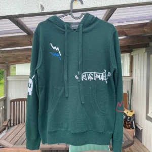 Mörkgrön hoodie från Zadig & Voltaire - Säljer en mörkgrön hoodie från Zadig & Voltaire i 100% kashmir. Tröjan har coola broderade detaljer med graffiti-inspirerade motiv i vitt, blått och rött, samt snörning vid huvan och känguruficka framtill. Perfekt för dig som gillar streetstyle och mjuka material.