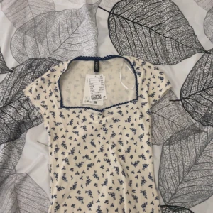 Blommig topp med blå spetskant hm - Supersöt topp från hm i ljusbeige med små blå blommor. Toppen har korta ärmar, fyrkantig ringning och blå spetskant runt halsen. Aldrig använt