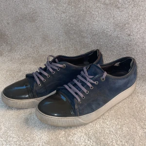 Blå sneakers från Lanvin med lackad tå - Snygga blå sneakers från Lanvin med cool lackad svart tå och matchande blå mocka på sidorna. Skorna har lila snörning och vit platt sula. Perfekt för dig som gillar stilrena detaljer och vill sticka ut med en unik kombo av material. Priset är ej hugget i sten