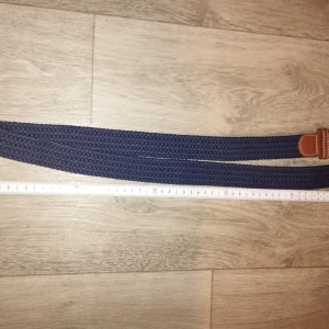 Mörkblå flätad bälte med skinn - Snyggt mörkblått flätat bälte med detaljer i brunt skinn och en stilren silverfärgad metallspänne. Bältet har en klassisk look med kontrastsömmar på skinnet och passar perfekt till jeans eller chinos. Flätad design gör det enkelt att justera längden efter behov.