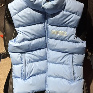Ljusblå dunväst från Hoodrich - Snygg ljusblå dunväst från Hoodrich i storlek M. Västen har ett quiltat mönster, hög krage och två sidofickor. Stängs med dragkedja framtill och har Hoodrich-loggan tryckt på bröstet. Perfekt för lager-på-lager och streetwear. Den har låte smuts men går att rensa bort har används max 5ggr köptes några år sedan men har aldrig använts.
