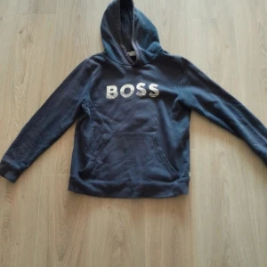 Mörkblå hoodie från BOSS - Snygg mörkblå hoodie från BOSS med vit logga på bröstet. Klassisk känguruficka framtill och stor huva. Tröjan har långa ärmar och är gjord i mjuk bomull, perfekt för chill dagar. Passar dig som gillar stilren streetwear och vill ha ett cleant plagg i garderoben. Tröjan har skavanker på armbågarna men man tänker inte på det, Va inte rädda om att fråga om att ändra på priset för då försöker vi lösa det!🫶