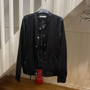 Svart bomberjacka från Daphnea Paris - Svart bomberjacka med silkeslen finish från Daphnea Paris. Jackan har snygga silverfärgade knappar framtill och cool broderad design med två fåglar på ryggen. Långärmad och med ribbade muddar för en chill vibe. Perfekt för dig som gillar detaljer och stil. Frakt ej inklusive.