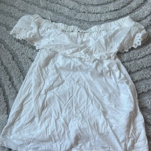 Vit offshoulder topp med spets från Yessica - Superfin vit offshoulder topp från Yessica i mjuk bomull. Toppen har bred spetsvolang med broderade detaljer längs axlarna och elastisk kant som håller den på plats. Perfekt för sommarens chill dagar och har en luftig, loose passform.