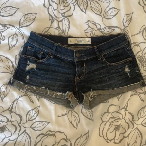 Mörkblå jeansshorts Abercrombie & Fitch - Snygga mörkblå jeansshorts från Abercrombie & Fitch med slitna detaljer och fransig kant. Klassisk femficksmodell med låg midja och kontrastsömmar. Perfekt för dig som gillar en chill och avslappnad stil på sommaren.