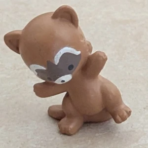 Ett litet plast djur - En liten brun figur som liknar ett djur, troligen en tvättbjörn eller björn, med vit och mörkbrun ansiktsmålning. 