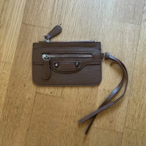 Brun plånbok med dragkedja - Snygg brun plånbok i skinn med dragkedja och handledsrem. Perfekt för att hålla dina kort och kontanter säkra. Den har en stilren design med silverdetaljer och knappar.