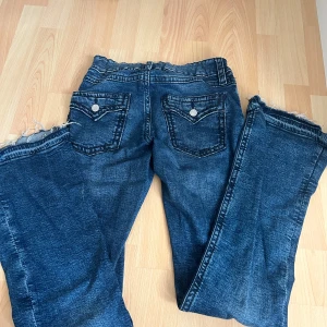 Blå jeansbyxor Y2k - Snygga blå jeansbyxor  med coola bakfickor med knappdetaljer och råa, fransiga benslut. Byxorna har en elastisk midja för extra komfort och en klassisk tvättad look som passar till det mesta. Perfekt för dig som gillar avslappnad stil.