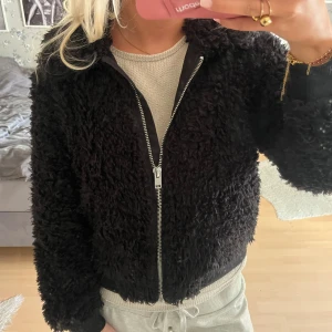 Svart teddyjacka med dragkedja - Mysig svart teddyjacka med fluffig struktur och hel dragkedja framtill. Jackan har ribbade muddar vid ärmslut och midja för en skön passform. Perfekt för dig som vill ha en trendig och bekväm look under kyligare dagar.