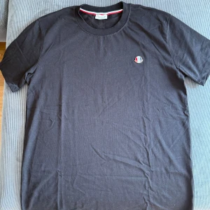 Svart t-shirt från Moncler - Svart t-shirt från Moncler med rund hals och korta ärmar. Diskret Moncler-logga på bröstet och en snygg röd-vit-blå detalj på insidan av kragen. Mjuk bomullskvalitet som känns skön mot huden. Perfekt för en clean och stilren look.