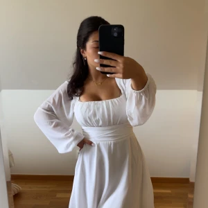 Vit omlottklänning med puffärmar - Superfin vit klänning med omlottband i midjan och puffiga långärmar i transparent tyg. Klänningen har en fyrkantig urringning och markerad midja, vilket ger en snygg siluett. Perfekt för sommarens alla tillfällen och har en flowy känsla. Har även en rosset där bak. Använd en gång.