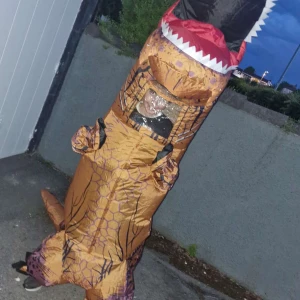 Uppblåsbar T-Rex dräkt brun/orange - Säljer en uppblåsbar T-Rex dinosauriedräkt i brun och orange med svarta ränder och detaljer. Dräkten har genomskinligt fönster vid ansiktet, inbyggd fläkt och USB-batteripack för att hålla den uppblåst. Perfekt för att sticka ut på temakvällar eller maskerad! Köpte bästa kvalité och sen tog jag på mig den 2 gånger till skolan i spex och nu säljer jag den som ny.