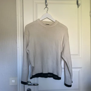 Beige stickad tröja med svarta detaljer - Säljer en beige stickad tröja med svarta ribbade kanter vid ärmslut och nederkant. Tröjan har en rund hals och långärmade, lite loose fit. Perfekt för dig som gillar enkel men snygg stil och vill ha något som funkar till jeans eller kjol.