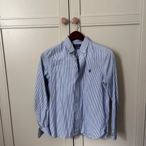 Blårandig skjorta från Polo Ralph Lauren - Snygg blårandig skjorta från Polo Ralph Lauren i slim fit-modell. Skjortan har klassisk krage, knappar framtill och lång ärm med knapp vid manschetten. Tillverkad i mjuk bomull och har den ikoniska Polo-loggan broderad på bröstet. Perfekt för en clean och fräsch look.
