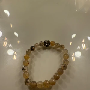 Beige pärlarmband med lejonhuvud - Snyggt armband med runda pärlor i beige och grå toner, dekorerat med ett coolt lejonhuvud i silverfärg. Pärlorna har ett unikt mönster med mörka inslag som gör varje pärla speciell. Perfekt accessoar för dig som vill sticka ut med något annorlunda.