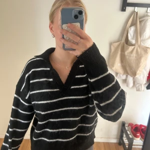 Svartvitrandig stickad tröja med krage - Mysig svart stickad tröja med vita horisontella ränder och v-ringad hals. Tröjan har en bred krage och långa ärmar. Passformen är loose och materialet känns mjukt och fluffigt.