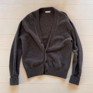 Mörkgrå kofta från Mango, storlek M - Säljer en stilren mörkgrå kofta från Mango i herrstorlek M. Koftan har djup v-ringning, ribbade muddar och knäppning med tre svarta knappar framtill. Materialet är mjukt och stickat, perfekt för lager-på-lager under kyliga dagar. Lite nopprig har den blivit av användning, men fortfarande bra skick.